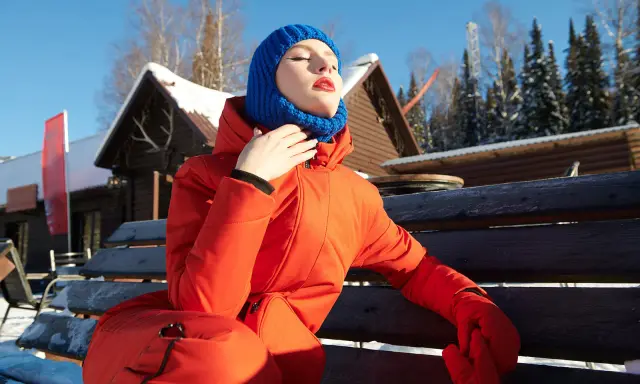 Kobieta w pomarańczowej kurtce i niebieskiej balaklawie, co to za ochrona przed zimnem, siedzi na ławce na tle zaśnieżonych domków i lasu.