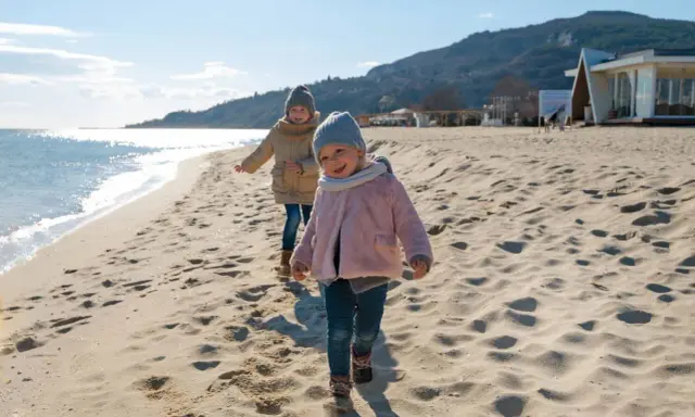 Dwa uśmiechnięte dzieci biegają po piaszczystej plaży. Słońce odbija się od wody, a w tle widać wzgórza i budynek.