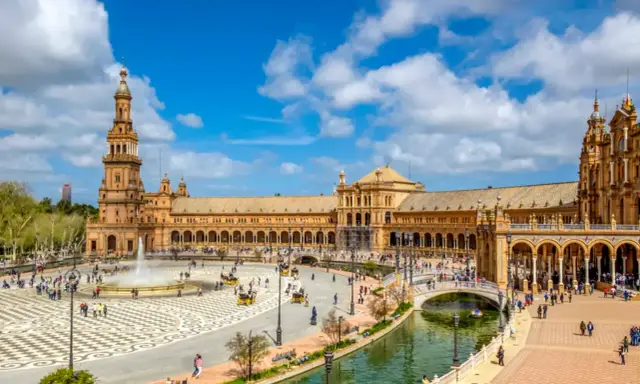 Der beeindruckende Sevilla Palast mit seinen Türmen und Arkaden, umgeben von einem Kanal und belebtem Platz mit Brunnen und Kutschen.