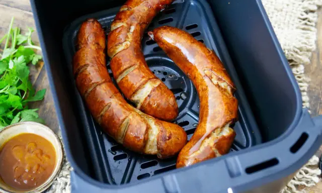 Drei frische grobe Bratwürste garen in einem Airfryer. Daneben eine Schale mit Dip und Petersilie.