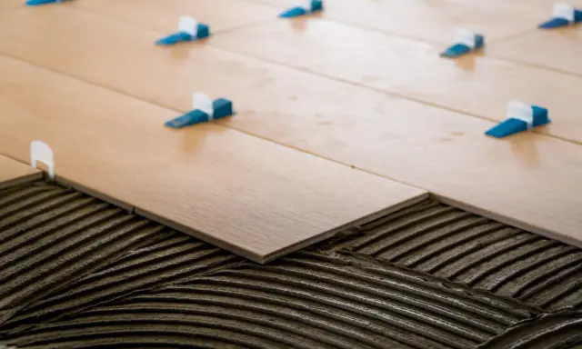 Pose de carrelage 60x60 en cours. Des carreaux en bois clair sont posés sur du mortier frais, avec des croisillons bleus et blancs pour un espacement parfait.