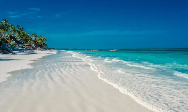 En vit sandstrand med turkost vatten och palmer vid Kendwa, Zanzibar. Båtar guppar i fjärran.