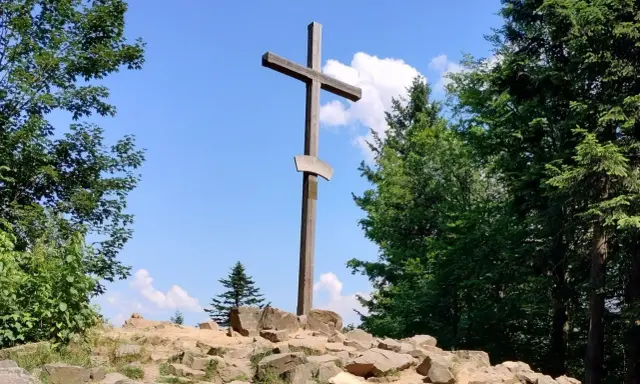 Drewniany krzyż na skalistym szczycie w Górach Świętokrzyskich. Otaczają go bujne drzewa i błękitne niebo z chmurami.