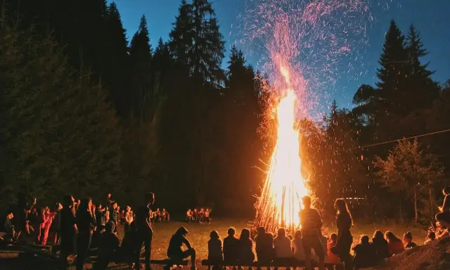 Människor samlas runt en stor brasa i skogen, en vacker svensk tradition som lyser upp natten med gnistor och värme.
