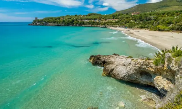 Spiaggia dorata e mare cristallino nel Cilento, con una torre antica su una scogliera e colline verdi sullo sfondo.