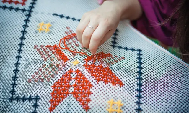 Dłoń z igłą i czerwoną nitką wyszywa wzór na białej kanwie. Widać fragmenty czerwonych i żółtych gwiazdek. Zamek otwiera się w kierunku ręki.