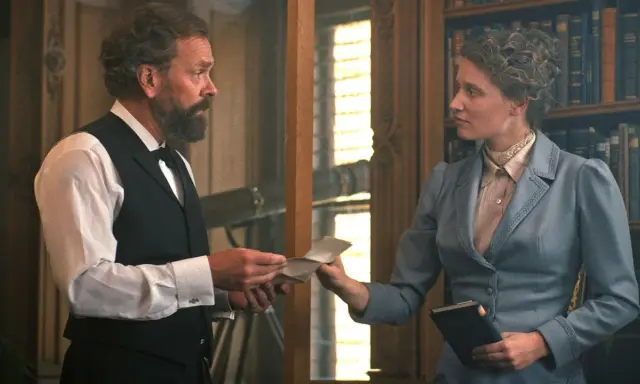 En scen från en alfred nobel film. En man med skägg ger ett brev till en kvinna i en blå dräkt framför en bokhylla.