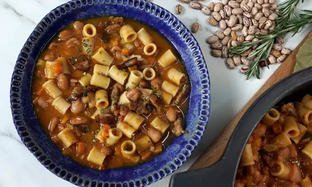 Un reconfortante plato de pasta e fagioli en un cuenco azul, con frijoles y romero esparcidos alrededor.