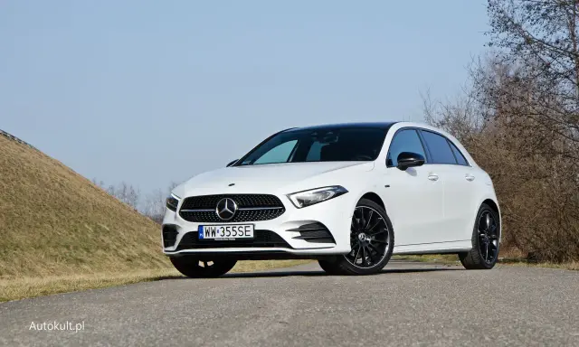 Biały Mercedes A250 z czarnym dachem i felgami stoi na drodze, na tle wzgórza i drzew.