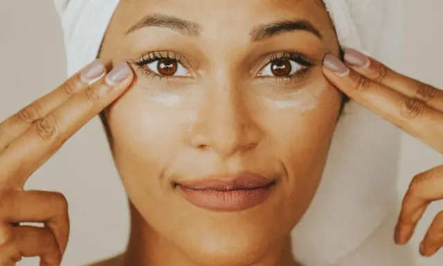 Une femme applique de la crème contour des yeux sur tout le visage, avec une serviette sur la tête.