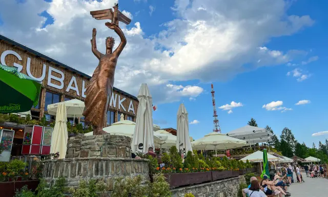 Gubałówka atrakcje: pomnik z orłem, restauracje, parasole i ludzie odpoczywający na ławkach.