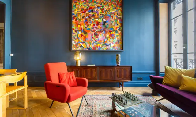 Ein Wohnzimmer mit einem farbenfrohen Gemälde im art déco zeitraum, rotem Sessel, Holzsideboard und einem Schachspiel auf dem Couchtisch.