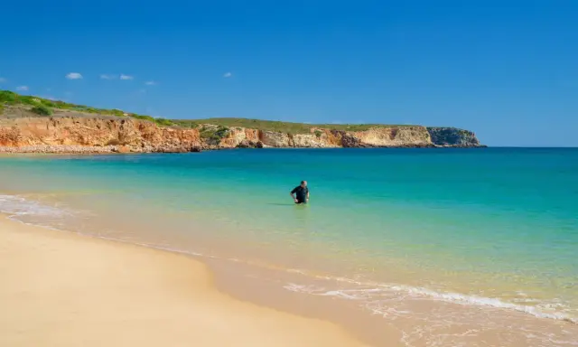 Ein Mann im Neoprenanzug steht im türkisfarbenen Wasser am Praia do Martinhal. Im Hintergrund ragen ockerfarbene Klippen auf.