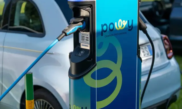 Estación de recarga para coche eléctrico con cable azul conectado. La estación tiene el logo "powy" y el lema "CHARGE THE CHANGE".