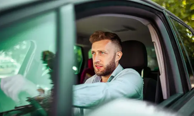 Mężczyzna z zarostem, w jasnej koszuli, siedzi za kierownicą. Jego profil sugeruje skupienie, jakby wypełniał profil kierowcy wniosek.