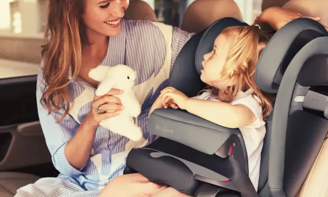 Uśmiechnięta mama z pluszowym zajączkiem przytula córkę w foteliku samochodowym Cybex Pallas 9-36 kg.