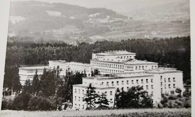 Duży budynek sanatorium w otoczeniu drzew, na tle pagórkowatego krajobrazu.