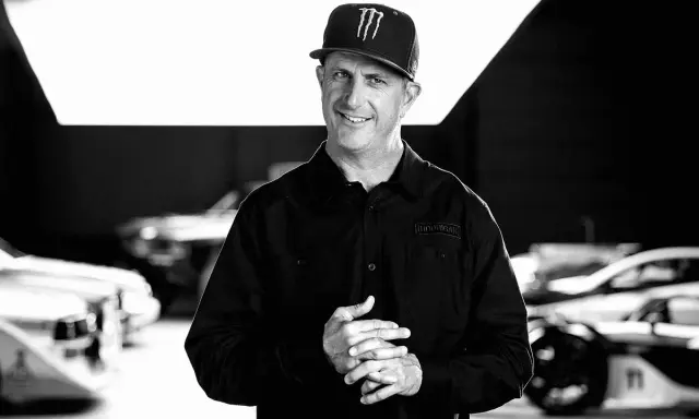 Ken Block, sonriente y con gorra, posa frente a coches de rally.