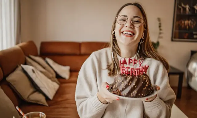 Vrolijke vrouw met bril en koptelefoon viert haar verjaardag met een chocoladetaart. Ze heeft de volwassen leeftijd bereikt en straalt van geluk.