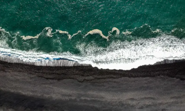 Gran Canaria: Türkisfarbenes Meer trifft auf einen dramatischen schwarzen Strand. Wellen brechen sich auf dem dunklen Sand.