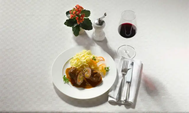 Ein Teller Rouladen mit Bandnudeln und einem Glas feinherb Wein. Dazu ein kleines Blümchen und Besteck.