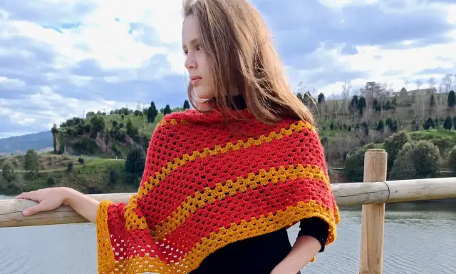 Chica con un pañuelo triangular crochet rojo y amarillo, apoyada en una barandilla de madera junto a un lago.