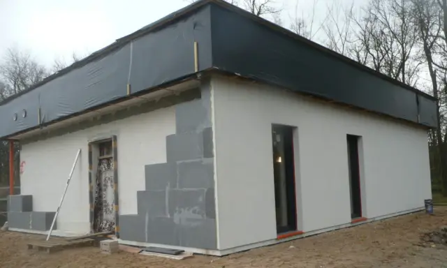 Budowa domu ze styropianu w toku. Ściany z bloczków, częściowo otynkowane, dach przykryty czarną folią.