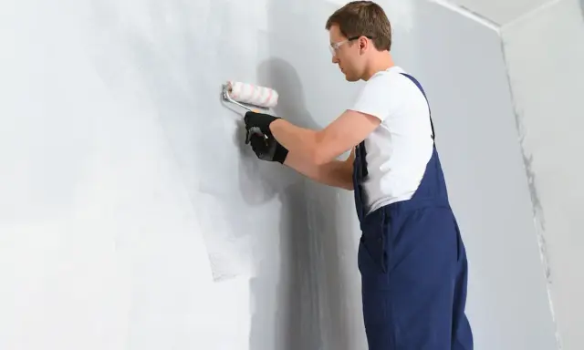 Mężczyzna w ogrodniczkach maluje ścianę wałkiem. Nakłada nową farbę na starą, tworząc gładką powierzchnię.