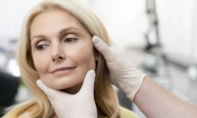 Kobieta po 50. roku życia podczas konsultacji dotyczącej zabiegów na twarz. Dłonie w rękawiczkach delikatnie dotykają jej policzków.