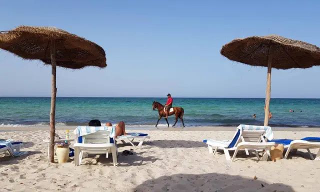 Piękna plaża na Djerbie, Tunezja. Jeździec na koniu przemierza brzeg morza, podczas gdy inni odpoczywają pod parasolami.