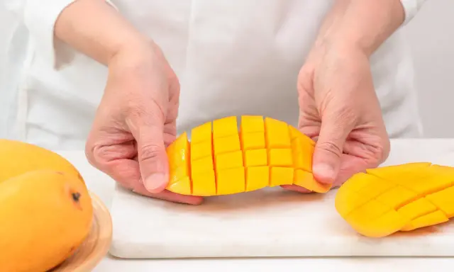 Ręce trzymają pokrojone w kostkę mango, pokazując, jak pokroić mango. Obok leżą całe owoce.