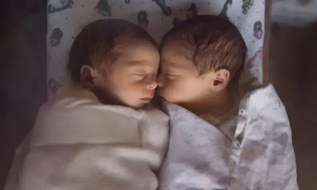Dwa śpiące niemowlaki, każde w wieku około 2 miesięczne, wtulone w siebie pod białymi kocykami.