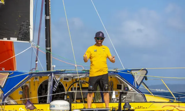 Arnaud Boissières, souriant, fait un double pouce levé sur son voilier jaune et bleu, prêt pour une nouvelle aventure en mer.