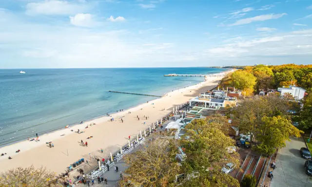 Słoneczna kołobrzeg plaża z ludźmi spacerującymi i odpoczywającymi. W tle morze i molo, a po prawej stronie drzewa w jesiennych barwach.