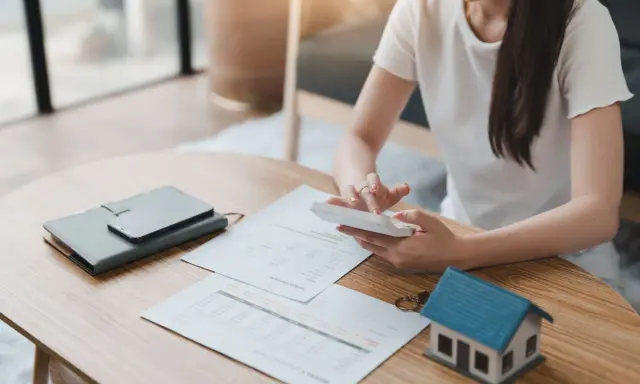 Persona calculando gastos en una mesa con un modelo de casa, documentos y calculadora. Se pregunta quién paga el alta de suministros en un alquiler.