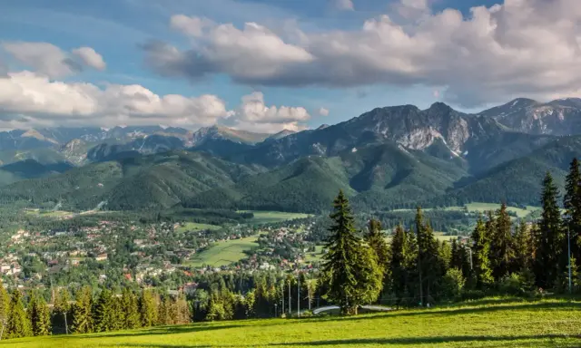 Zielona polana z widokiem na malownicze lasy w Tatrach, miasteczko i skaliste szczyty w tle.