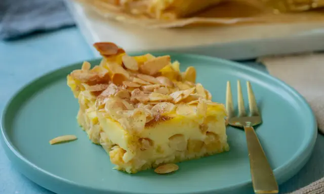 Ein Stück Apfel Schmand Kuchen mit Mandelblättchen bestreut, auf einem türkisfarbenen Teller, daneben eine goldene Gabel.