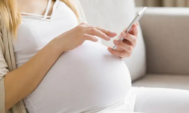 Kobieta w ciąży, w białej bluzce i kardiganie, siedzi na kanapie i korzysta z telefonu. Może sprawdzać zwolnienie lekarskie w ciąży.