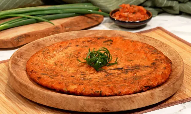 Ein goldbrauner, herzhafter Kimchi-Pfannkuchen, garniert mit Frühlingszwiebeln, liegt auf einem Holzteller. Daneben eine Schale mit Kimchi und grüne Frühlingszwiebeln.
