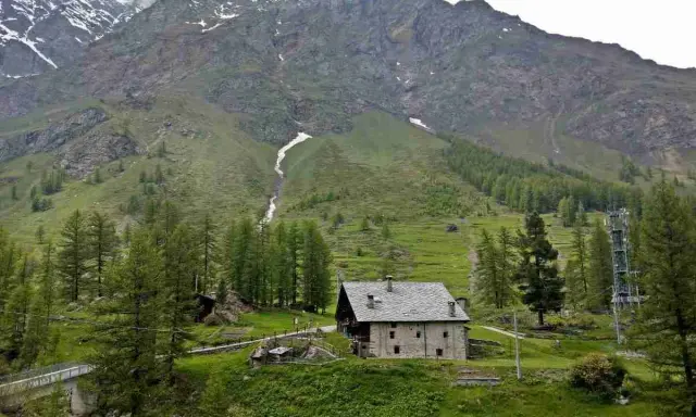 Un casolare in pietra nel **Val di Rhemes**, circondato da verdi pendii e imponenti montagne innevate.