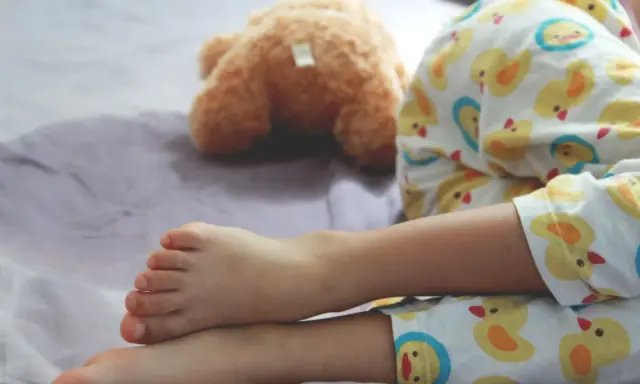 Nocne wpadki zdarzają się nawet 5-latkom. Dziecko w piżamce z kaczuszkami śpi obok pluszowego misia, a na pościeli widać mokrą plamę.