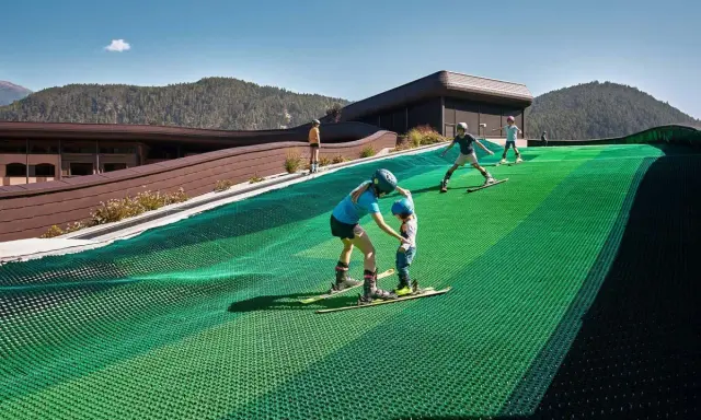 Bambini imparano a sciare su una pista verde al Falkensteiner Family Resort Lido, con montagne sullo sfondo.