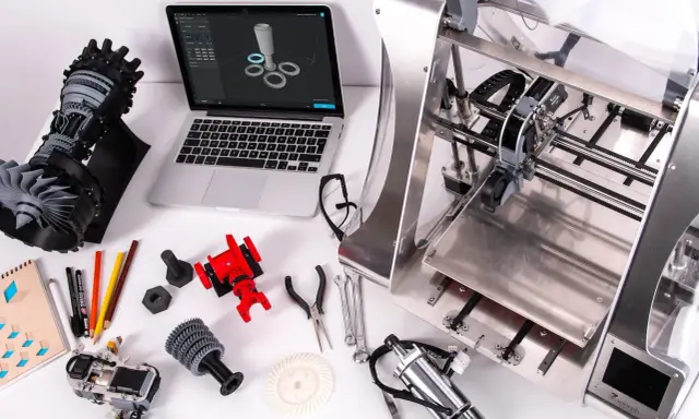 Il disegnatore CAD lavora su un laptop con un modello 3D, circondato da componenti stampati in 3D e strumenti.