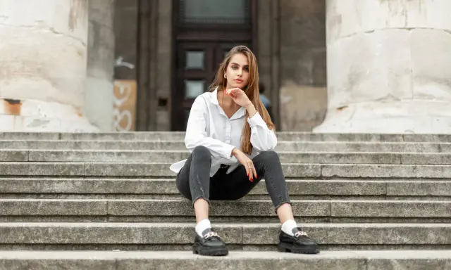 Eleganckie mokasyny to idealne buty do szarych spodni. Kobieta w białej koszuli i czarnych jeansach siedzi na schodach.