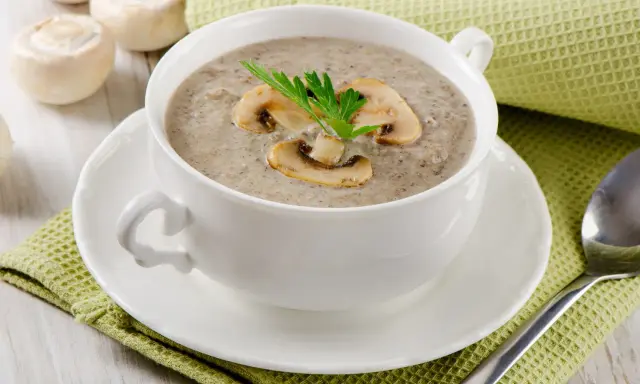 Un réconfortant velouté de champignon, garni de lamelles et d'une touche de persil, servi dans une tasse blanche sur une serviette verte.