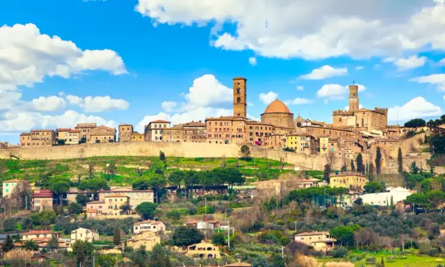 Panoramica di Volterra, dove si trova questo antico borgo toscano, con le sue torri e case arroccate sulla collina sotto un cielo azzurro con nuvole bianche.