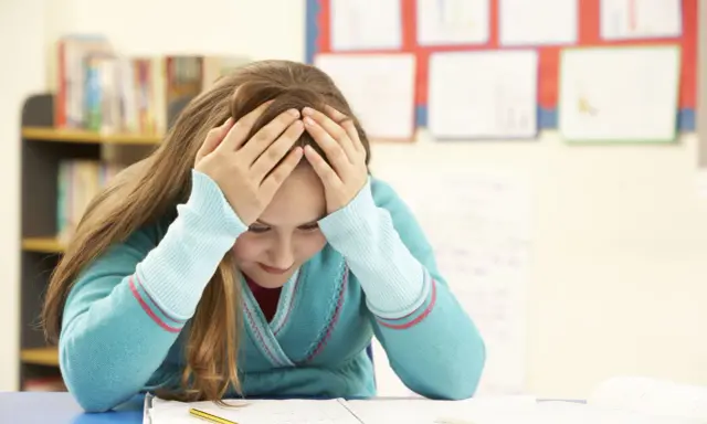 Verzweifeltes Mädchen mit Händen am Kopf im Klassenzimmer. Die **Kollektivstrafe Schule** lastet schwer auf ihr.
