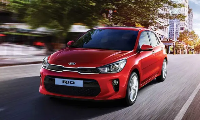 Un hatchback rojo Kia Rio se desplaza por una calle urbana, con edificios y árboles borrosos por el movimiento.