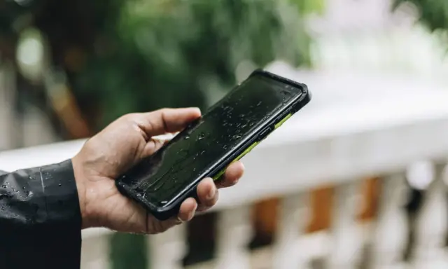 Dłoń trzyma mokry smartfon ip67 na tle zieleni. Krople deszczu spływają po ekranie.