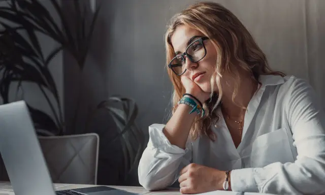 Zamyślona kobieta przy laptopie, z podpartą głową na dłoni. Wokół niej panuje atmosfera rezygnacji, jakby mówiła sobie: "nie mam marzeń".